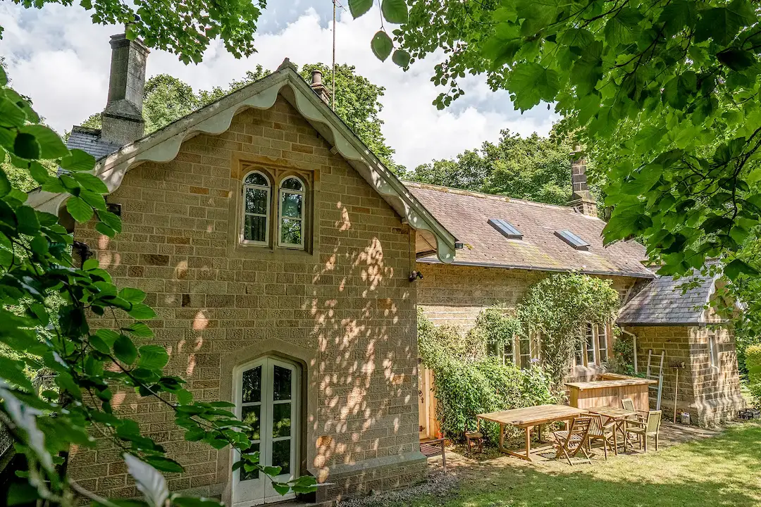 A stone cottage with a sloped roof, surrounded by greenery, featuring windows and a wooden outdoor table with chairs.