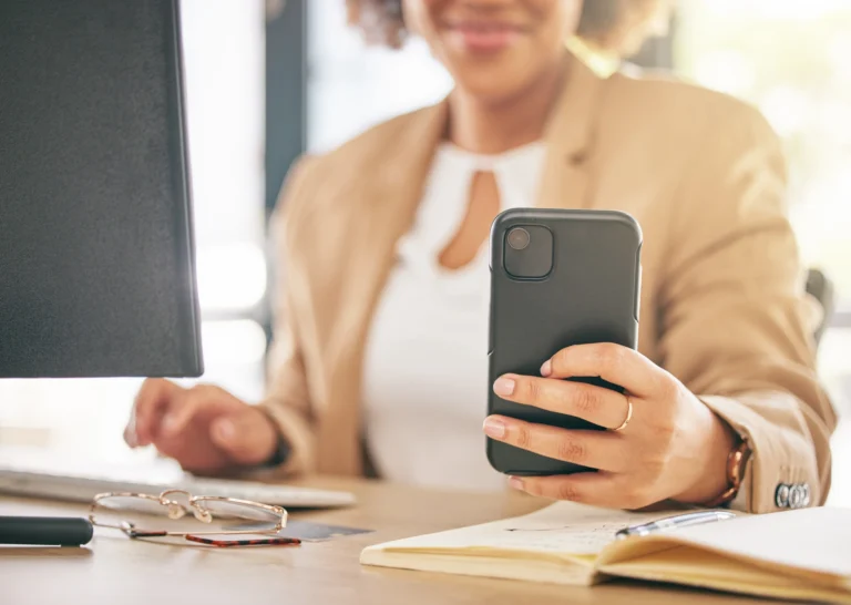Woman’s hands using a smartphone and laptop on a desk with a notepad and glasses, illustrating digital marketing and multitasking work.